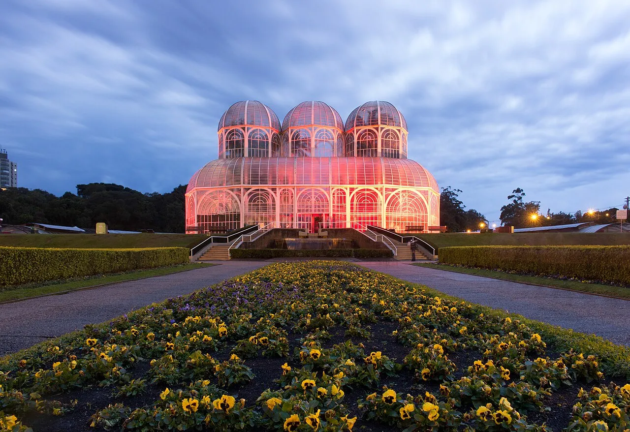 Estufa de vidro do Jardim Botânico de Curitiba iluminada em tons de rosa ao anoitecer, com canteiro de flores amarelas em primeiro plano