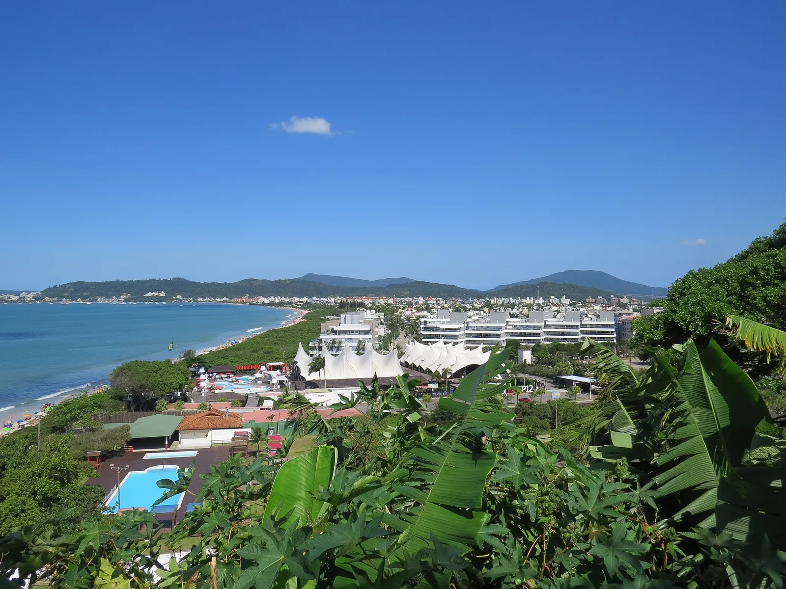 Vista do alto de Jurerê Internacional, em Florianópolis, mostrando a faixa de areia, os beach clubs de lona branca à beira-mar e os condomínios horizontais de baixa altura do bairro