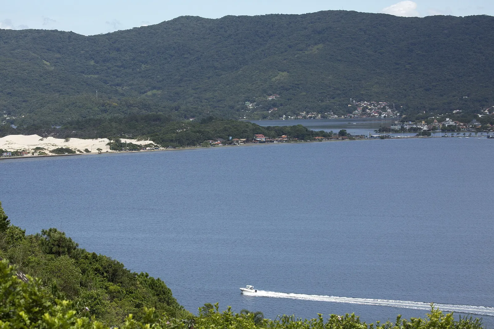 Vista do alto da Lagoa da Conceição, em Florianópolis, com a lagoa em primeiro plano, as dunas da Joaquina à esquerda e casas de baixa altura espalhadas pela encosta ao fundo