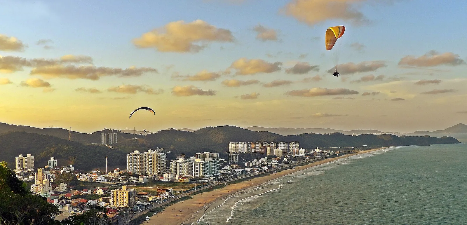 Praia Brava, em Itajaí, vista do alto ao entardecer, com a faixa de areia curva, a avenida beira-mar e os prédios residenciais alinhados à orla, além de parapentes no céu