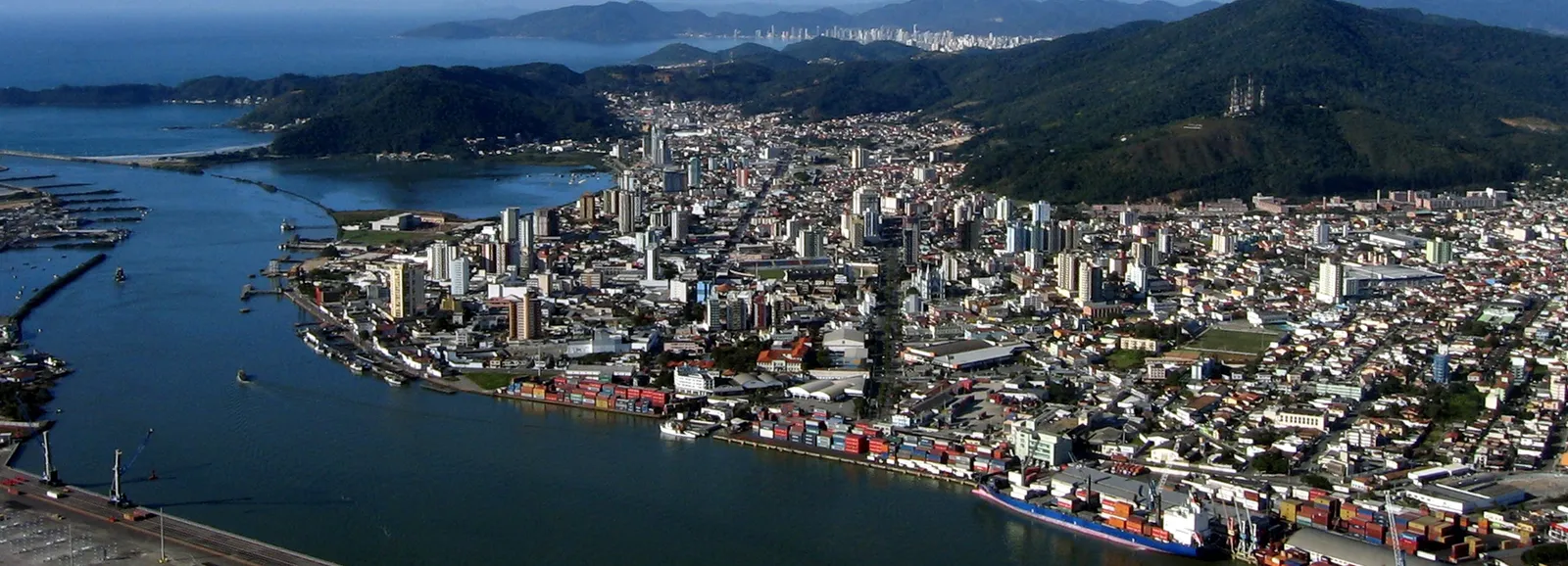 Vista aérea de Itajaí mostrando o terminal de contêineres às margens do rio Itajaí-Açu, os prédios do centro da cidade logo atrás e a silhueta dos arranha-céus de Balneário Camboriú ao fundo