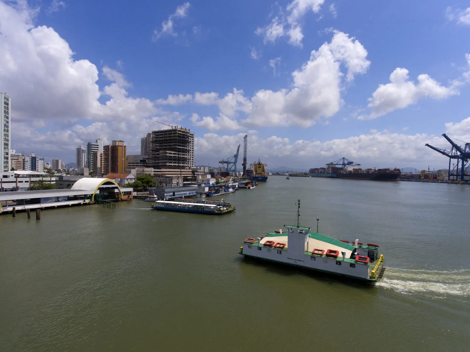 Ferry-boat da travessia Itajaí-Navegantes cruzando o rio Itajaí-Açu, com outra embarcação ao fundo, prédios do centro urbano e guindastes do porto na margem