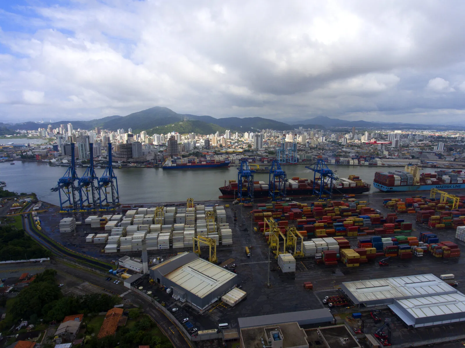 Vista aérea do complexo portuário de Itajaí e Navegantes, com guindastes, contêineres empilhados e navios cargueiros nas margens do rio Itajaí-Açu, com o centro urbano e morros ao fundo