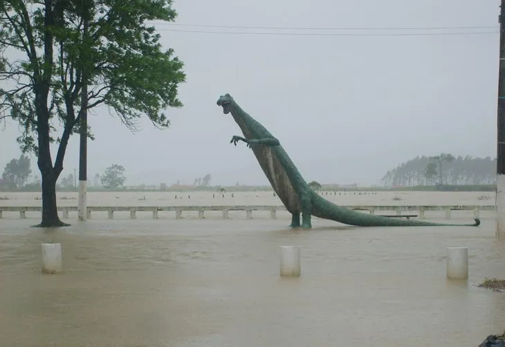Estátua de dinossauro, marco visual às margens da BR-101 em Tijucas, cercada por água barrenta durante uma enchente do rio Tijucas